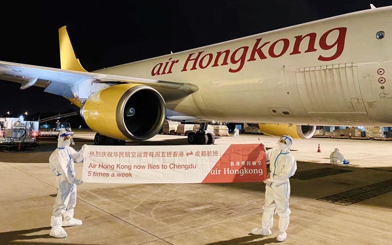 Two people dressed up in personal protective equipment holding up a banner in front of an Air Hong Kong plane